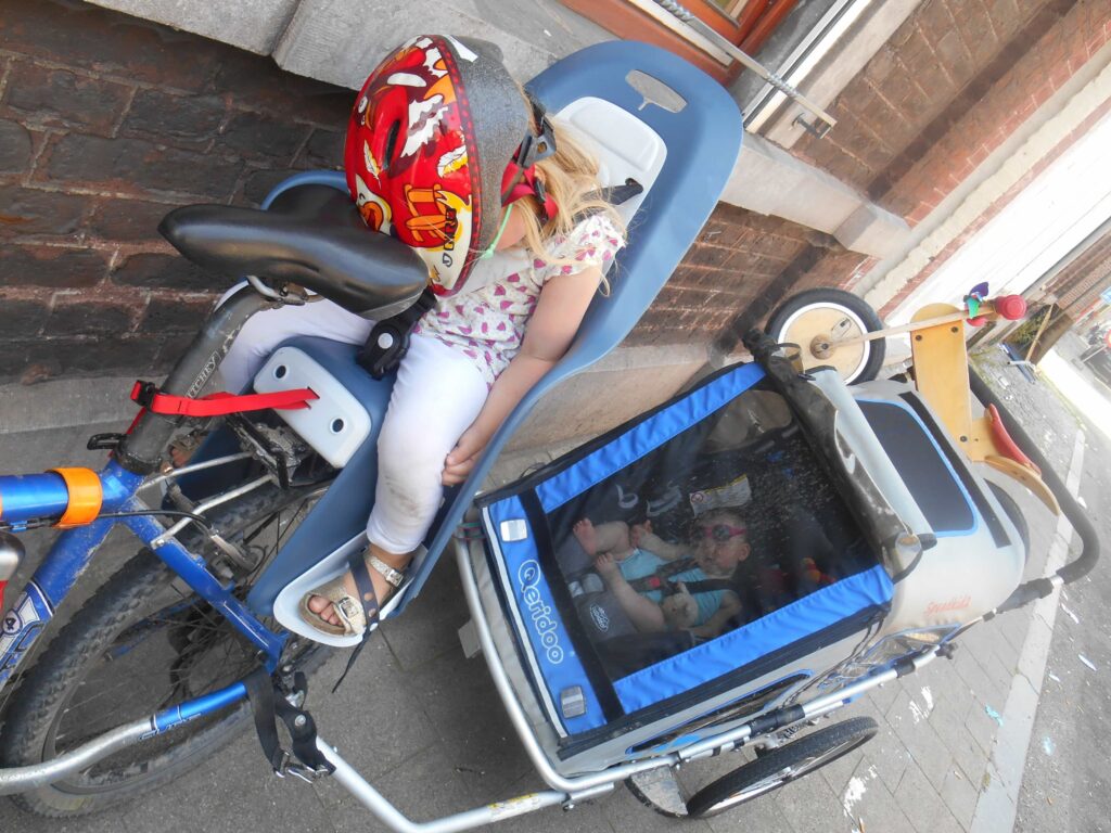 Transporter les enfants de 9 mois à 5 ans.
Photo de la grande sur le siège enfant et la petite dans la remorque avec le maxicosy.