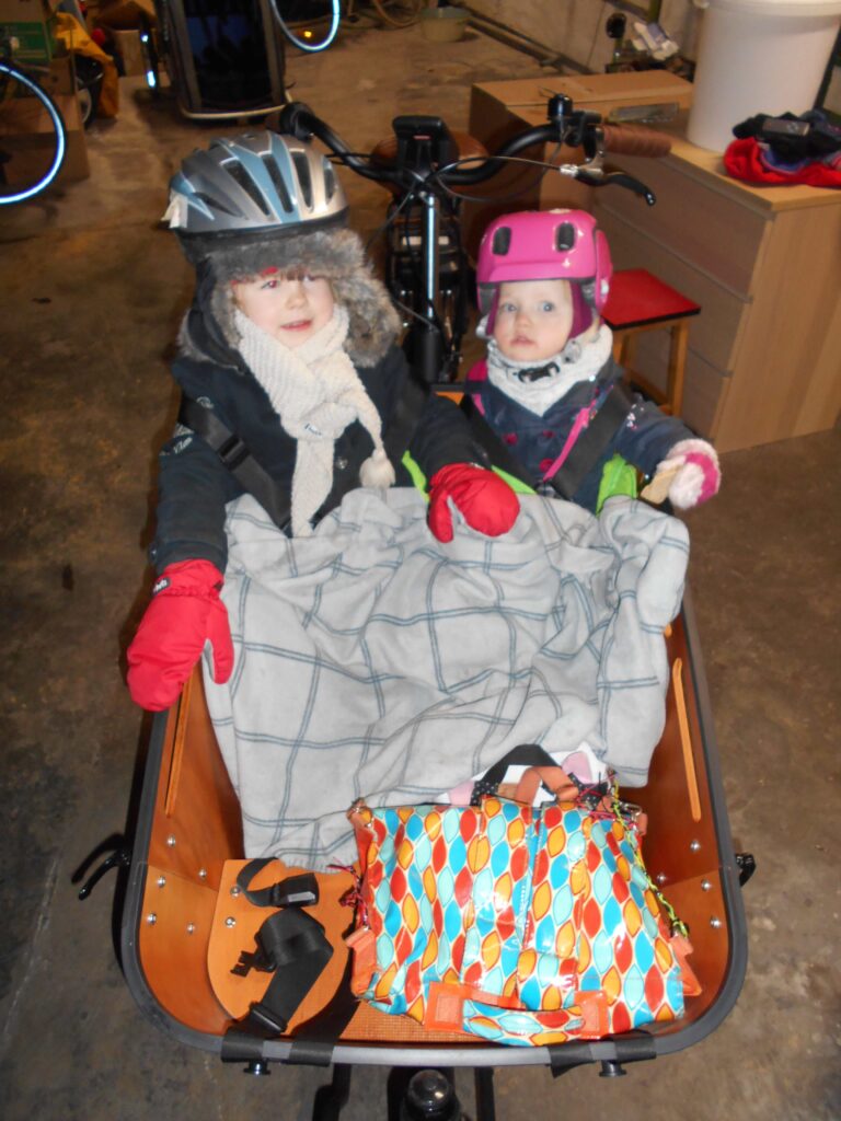 Storytelling du quotidien à vélo en famille.
Photo de mes filles assises dans le vélo cargo et équipées pour l'hiver...